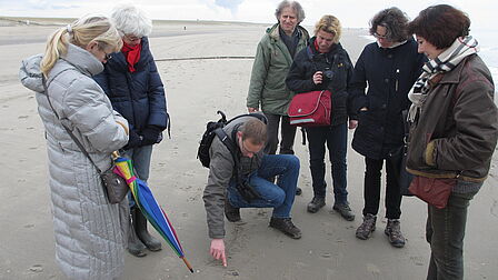Op de foto staan meerdere mensen in een kring om een man heen, die op de grond in het zand naar iets wijst.