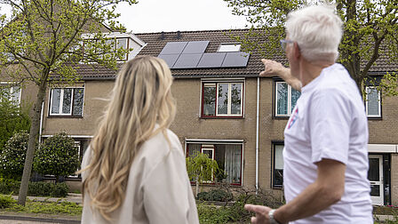Op de foto staan een vrouw en een man naast elkaar. De man wijst naar zonnepanelen die op het dak van een huis liggen. Verder zijn er twee bomen te zien.