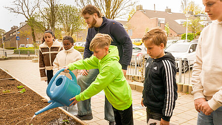 Op de foto zie je meerdere kinderen en een man. Een jongen met een groene jas giet water uit een gieter in de moestuin,