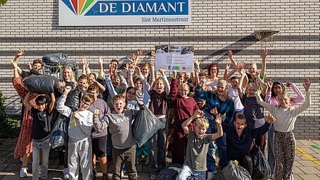Op de foto staan meerdere kinderen bij elkaar die een vuilniszak in de lucht houden of handen in de lucht steken. Tussen de kinderen staan meerdere volwassenen die meejuichen.