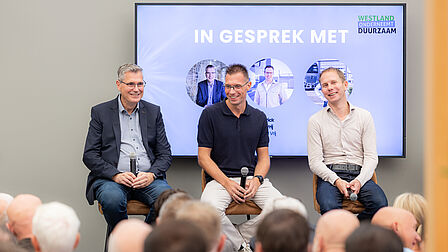 Op de foto is te zien dat drie mannen op een podium zitten en een menigte voor hen toespreken