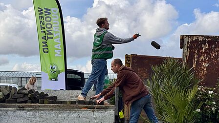 Op de foto zie je twee mannen en een vrouw op een terrein stenen verplaatsen. Er is ook een grote plant en een container te zien. De middelste persoon gooit met een steen.