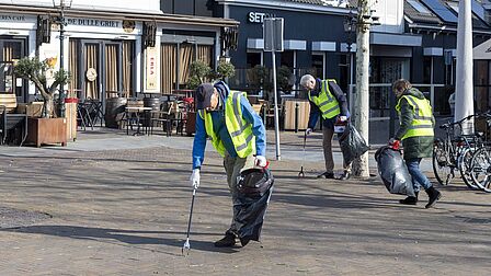 Op de foto rapen mensen met gele hesjes zwerfvuil van de straat op. Dat doen zij met een grijper.