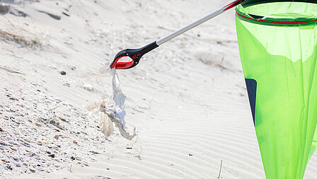 Op de foto is een groen zak en een grijper te zien. De grijper houdt een stuk afval vast. Op de achtergrond zie je zand van het strand.