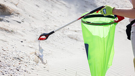Op de foto is een groen zak en een grijper te zien. De grijper houdt een stuk afval vast. Op de achtergrond zie je zand van het strand.