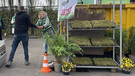 Op de foto neemt een vrouw van een man een tegel aan. Naast de vrouw staan meerdere plantenbakken met planten erin.